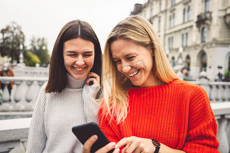 腰部以上的肖像中，两名穿着红色和米色毛衣的微笑的年轻女性在城市里走来走去，低头看着手机