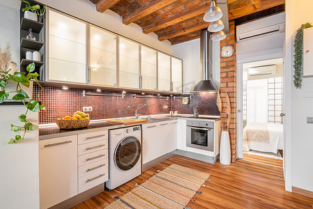 现代厨房内部配有木质天花板、优质电器炉灶和玻璃镶板壁柜，俯瞰着米色色调的卧室和砖玻璃墙。