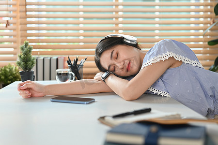 戴耳机的年轻漂亮亚洲女性睡着了，工作注意力分散，睡在办公桌前