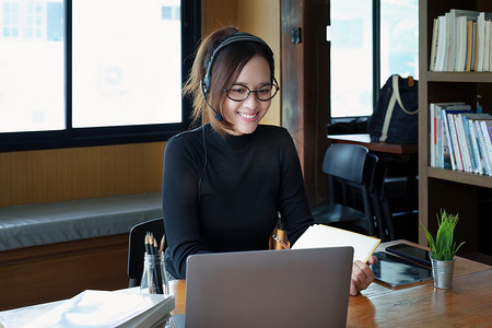 一位戴着眼镜、使用电脑笔记本电脑、耳机并使用笔记本电脑通过木制图书馆桌子上的视频会议在线学习的亚洲少女的肖像