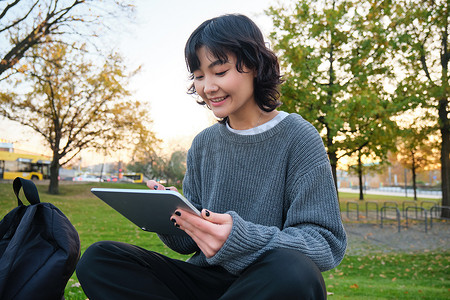 年轻微笑的韩国女孩、平面设计师、艺术家用钢笔工具在数字平板电脑上绘画的肖像，坐在公园里呼吸新鲜空气，抓耳挠腮，记笔记