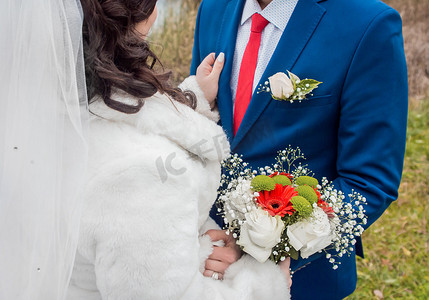身着白色大衣、头戴面纱的新娘站在身穿蓝色西装、红色领带、白色衬衫、手捧婚礼花束的新郎对面。