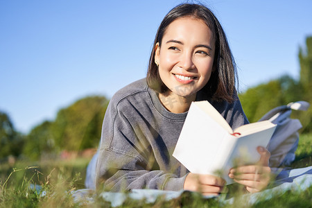 可爱的韩国女孩的肖像，在公园里躺在草地上读书，手里拿着最喜欢的书放松，开心地微笑