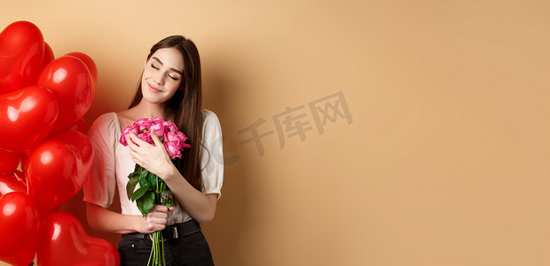 感性浪漫的女孩拥抱粉红玫瑰花束，闭着眼睛微笑，想着爱人，站在情人节心形气球附近，米色背景