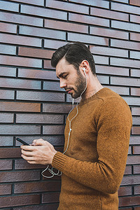 人气大牌摄影照片_微笑的男人戴着耳机听音乐，靠在砖墙上。