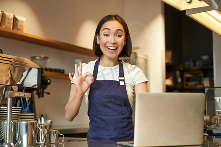 咖啡师女孩的肖像，亚洲女咖啡馆工人，显示好，好的标志批准，推荐某事，快乐和高兴，穿着围裙，站在柜台后面的笔记本电脑