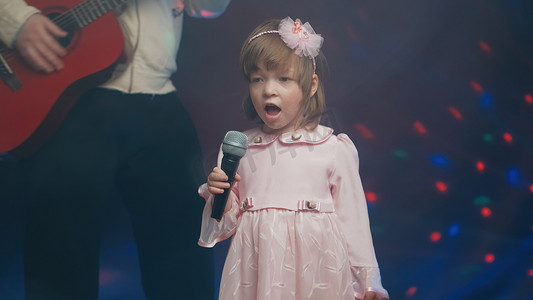 穿着复古连衣裙的小女孩在舞台上唱歌，她的父亲弹着原声吉他