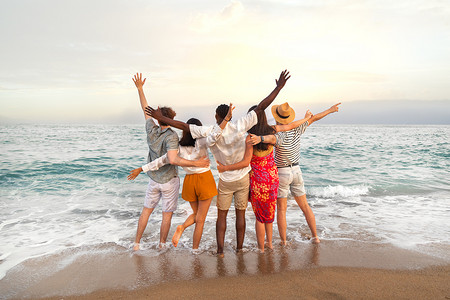 多种族朋友在度假旅行期间拥抱在一起，看着大海，举起双臂庆祝的后视图。