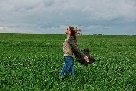 强风吹拂摄影照片_一位穿着外套的女人站在多云天气的田野中央，在强风中侧身转向镜头