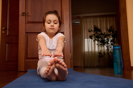 可爱的小女孩做伸展运动，赤脚坐在健身垫上，同时在室内锻炼和练习瑜伽