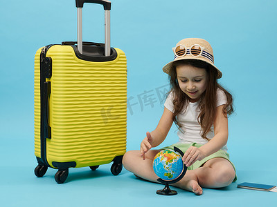 车票摄影照片_穿着夏装的可爱小女孩，坐在蓝色背景的黄色手提箱旁边，选择全球旅行目的地