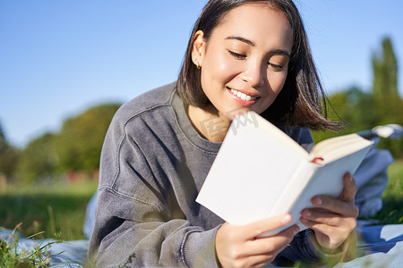 美丽微笑的亚洲女孩的肖像，在公园里读书，躺在草地上，手里拿着最喜欢的书。