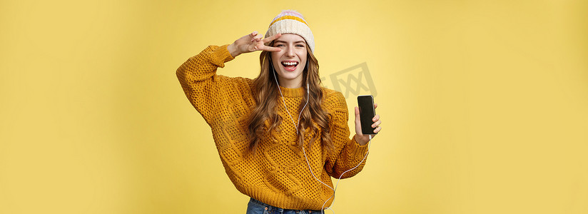 积极无忧无虑的迷人小女孩戴着有线耳机展示和平姿态，展示智能手机屏幕推广应用程序酷炫的全新手机，笑无忧无虑的黄色背景