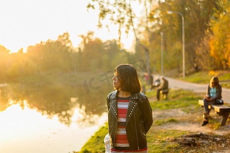 t恤深色摄影照片_穿着黑色夹克和条纹T恤的黑发女孩看着公园里的湖