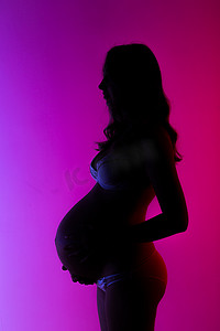 穿着内衣的美丽年轻孕妇在美丽的霓虹紫色灯光下，在工作室里愉快地抚摸着自己的肚子。