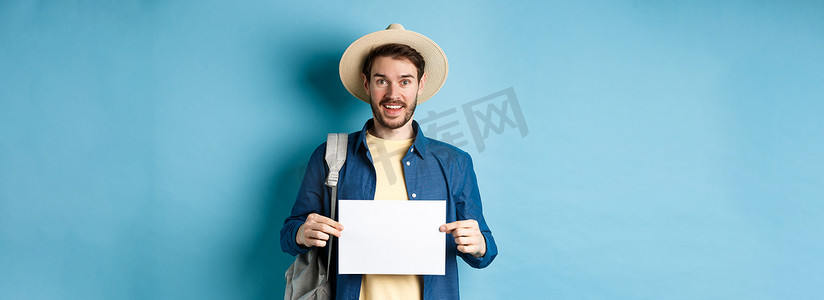 戴着夏帽的快乐游客，展示着一张空纸，微笑着，背着背包搭便车，站在蓝色背景上
