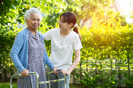 帮助器摄影照片_护理人员帮助亚洲老年女性患者在公园里用步行器行走，健康强大的医疗理念。