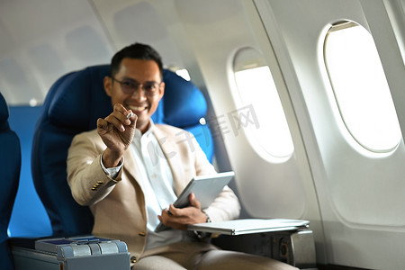 出差期间坐在飞机机舱中，穿着优雅豪华西装、使用数字平板电脑微笑的商人的形象