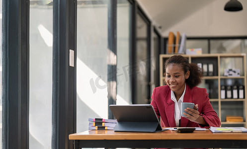 坐着的会计摄影照片_微笑的年轻非洲裔美国女性金融企业家坐在工作场所办公室，拿着咖啡杯，在笔记本电脑上工作，投资财务文件报告财务分析概念。