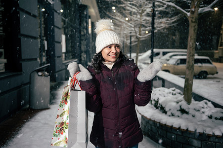 雪街摄影照片_带着购物袋的漂亮女人，沿着节日花环照亮的雪街漫步，捕捉雪花
