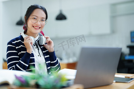 一名亚洲少女使用电脑、戴着耳机、使用笔记本通过图书馆木桌上的视频会议在线学习的肖像