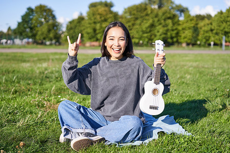 积极的韩国女孩坐在公园里，用手势展示尤克里里和摇滚，学习如何在户外演奏乐器