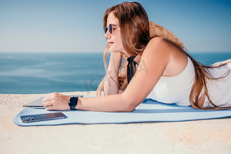 数字游牧者，戴帽子的女人，一位带着笔记本电脑的女商人在日落时坐在海边的岩石上，从远处进行在线商业交易。