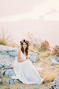 坐在山顶摄影照片_戴着花环的微笑新娘坐在山顶的石头上