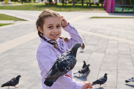 5岁快乐、可爱的白人小女孩微笑着看着镜头，一边喂手臂上的鸽子