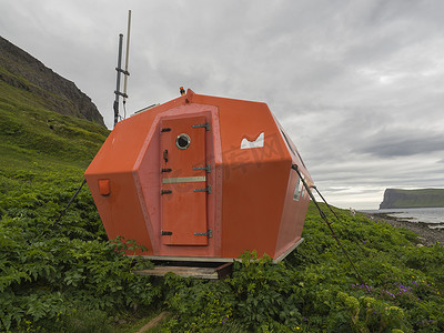 莫兰迪色调产品摄影照片_霍恩维克的红色紧急避难舱矗立在海滨草甸上，可欣赏冰岛西峡湾霍恩斯特兰迪尔霍恩比贾格悬崖的景色