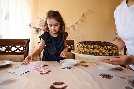 穿着时髦海军蓝裙子的迷人生日女孩玩着蜡烛，而她妈妈则把节日巧克力蛋糕放在桌上
