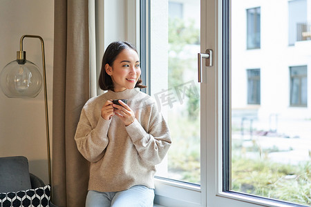 美丽的年轻亚洲女性坐在窗边喝咖啡，拿着浓缩咖啡杯，面带轻松、微笑的表情看着外面