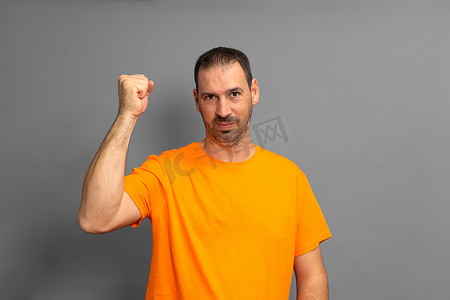 留着胡须的西班牙裔男子穿着橙色 T 恤，在灰色工作室背景下举起拳头庆祝某事。