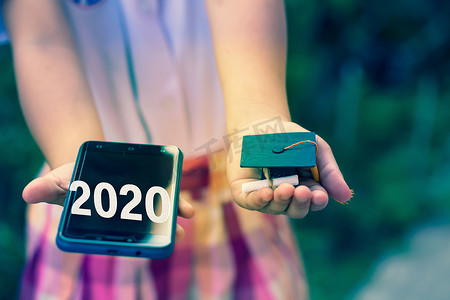 2020届毕业生和在线学习或电子学习概念：女学生伸出双臂，手上拿着智能手机和毕业帽。