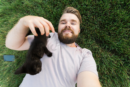 宠物主人摄影照片_男人用智能手机自拍，小猫躺在草地上玩耍 — 友谊、爱动物和宠物主人的概念