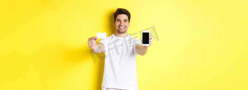 显示智能手机屏幕和信用卡、移动银行和网上购物概念、黄色背景的英俊白人男模特