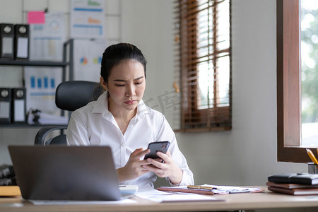 严肃的亚洲女商人使用笔记本电脑看着电脑打电话咨询客户坐在办公桌前，专注的女性销售经理拨打移动商务电话与办公室客户讨论工作