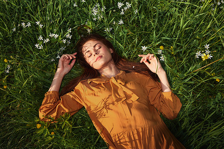 一个轻松的女人，躺在绿色的草地上休息，穿着橙色长裙，闭着眼睛，脸上挂着愉快的微笑，在夏日夕阳的温暖光芒照耀下恢复体力