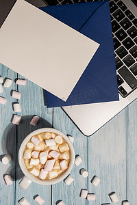 空贺卡或邀请卡模拟蓝色信封，在木蓝色桌子上放着白杯咖啡和棉花糖笔记本电脑键盘。