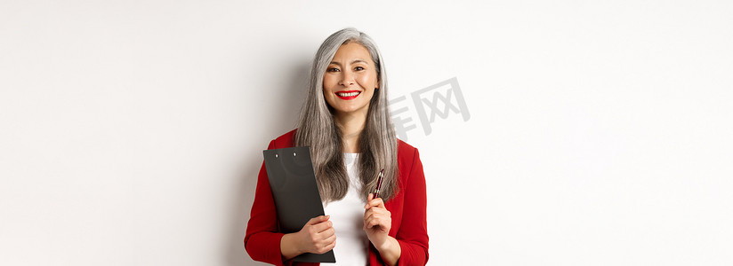 穿着红色西装外套的成功亚洲女老板，拿着带文件和笔的剪贴板，工作着，看起来很快乐，白色背景