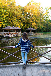 笼中女孩摄影照片_一位身穿格子衬衫的黑发年轻女子背靠池塘站立，享受秋日温暖的日子。