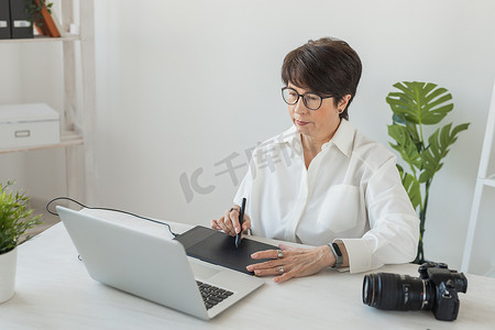 中年女性平面设计师或摄影师在现代办公室工作时使用数字图形平板电脑、坐在现代工作空间的专业女性修饰师、通用设计平板电脑和附近的相机