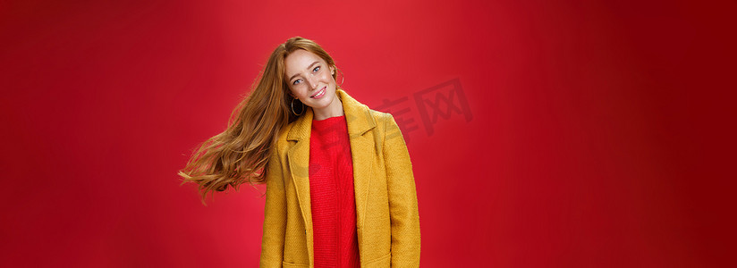 迷人的红发女性，长着雀斑，挥舞着头发，微笑着，头大倾，发型在空中飞扬，穿着温暖的黄色外套和针织连衣裙，在红色背景下摆出快乐和无忧无虑的姿势
