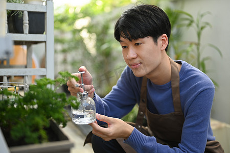 纯净水摄影照片_穿着围裙的亚洲男子满意地用喷雾瓶里的纯净水喷洒植物。