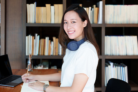 一名亚洲少女使用电脑和笔记本通过图书馆木桌上的视频会议在线学习的肖像