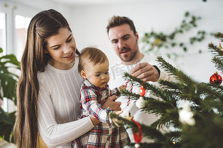 年轻快乐的三口之家带着女婴，装饰婴儿的第一棵圣诞树