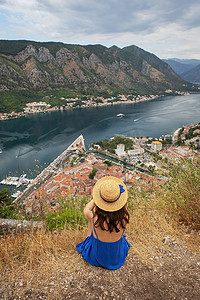 在黑山一个美丽的夏日，一个身穿蓝色连衣裙、头戴草帽的女孩坐在科托尔湾的背景下。