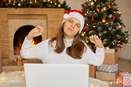 工作生活睡觉摄影照片_美丽的女人坐在地板上，在家里用笔记本电脑工作，围着圣诞树，伸着背，疲惫而放松，女孩穿着白色毛衣，戴着圣诞老人帽。