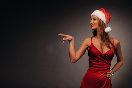 一位身穿红色连衣裙、头戴圣诞帽的女士用手指指着黑色背景。新年微笑的女孩在深色背景中展示方向