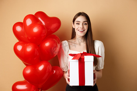 浪漫气球礼物摄影照片_浪漫的女孩站在心形气球旁，在情人节拿着惊喜礼物，对着镜头开心地微笑，站在米色背景中